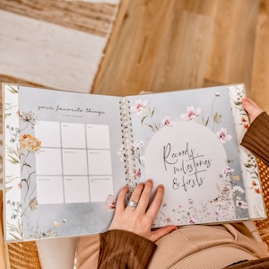 K&ouml;nnte beinhalten: Ein florales Baby-Erinnerungsbuch mit einer wei&szlig;en Seite, die zu einem Abschnitt mit dem Titel "Deine Lieblingsdinge mit einem Jahr" ge&ouml;ffnet ist. Die Seite hat ein Raster mit neun Quadraten, um Lieblingsdinge aufzuzeichnen. Die n&auml;chste Seite hat einen wei&szlig;en Kreis mit dem Text "Aufzeichnungen, Meilensteine & Erste Male" in Schwarz.