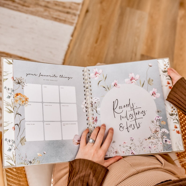 K&ouml;nnte beinhalten: Ein florales Baby-Erinnerungsbuch mit einer wei&szlig;en Seite, die zu einem Abschnitt mit dem Titel "Deine Lieblingsdinge mit einem Jahr" ge&ouml;ffnet ist. Die Seite hat ein Raster mit neun Quadraten, um Lieblingsdinge aufzuzeichnen. Die n&auml;chste Seite hat einen wei&szlig;en Kreis mit dem Text "Aufzeichnungen, Meilensteine & Erste Male" in Schwarz.