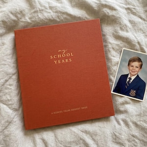 May include: A rust-coloured fabric-bound memory book with the gold-stamped text "my SCHOOL YEARS" and "A SCHOOL YEARS MEMORY BOOK" on the cover, accompanied by a rectangular photograph of a young person wearing a navy blue blazer and tie.