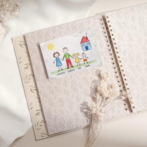 May include: An open spiral-bound journal with a light pink floral pattern. A child's drawing of a family and house is tucked inside. The drawing includes the words "MOMMY DADDY ME SAM".