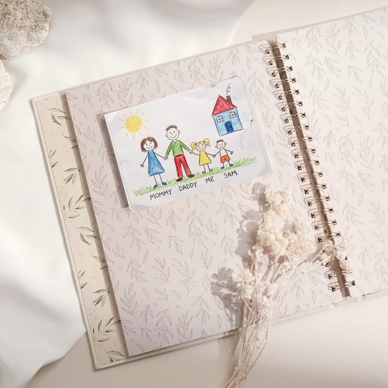 May include: An open spiral-bound journal with a light pink floral pattern. A child's drawing of a family and house is tucked inside. The drawing includes the words "MOMMY DADDY ME SAM".
