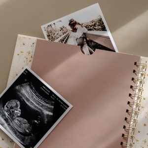 May include: A pink scrapbook with a gold spiral binding. The scrapbook has a floral pattern on the cover and is open to a blank page. There is a black and white ultrasound image and a photo of a couple on a wooden walkway in the background.