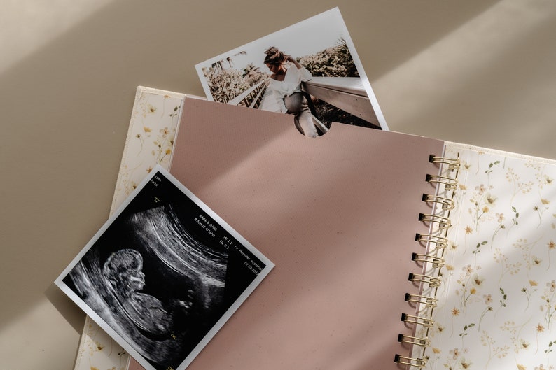 May include: A pink scrapbook with a gold spiral binding. The scrapbook has a floral pattern on the cover and is open to a blank page. There is a black and white ultrasound image and a photo of a couple on a wooden walkway in the background.