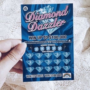 Pu&ograve; includere: Un biglietto della lotteria blu e rosa con le parole "Diamond Dazzler" e "Vinci fino a 100.000 &euro;". Il biglietto ha una griglia di diamanti con numeri da abbinare.