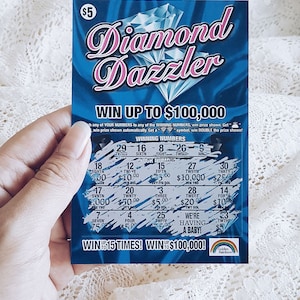 Pu&ograve; includere: Un biglietto della lotteria blu e rosa con le parole "Diamond Dazzler" e "Vinci fino a $ 100.000". Il biglietto ha un design a forma di diamante e un logo arcobaleno.
