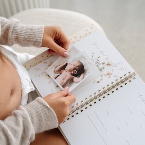May include: A white and gold baby memory book with a floral design. The book is open to a page that says "Welcome to the World" and has a photo of a newborn baby.