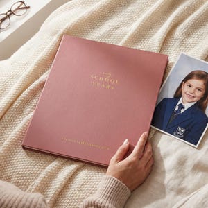 May include: A dusty rose-coloured memory book with gold foil text that reads My School Years and School Years Memory Book. The book is rectangular with a smooth cover. A photograph of a child in a navy blue blazer is placed next to the book.