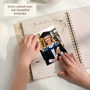 May include: A spiral-bound keepsake book with a photo of a graduate in a cap and gown. The book has the text "Graduation day" and "Every school year, one beautiful keepsake". The pages have a floral design.