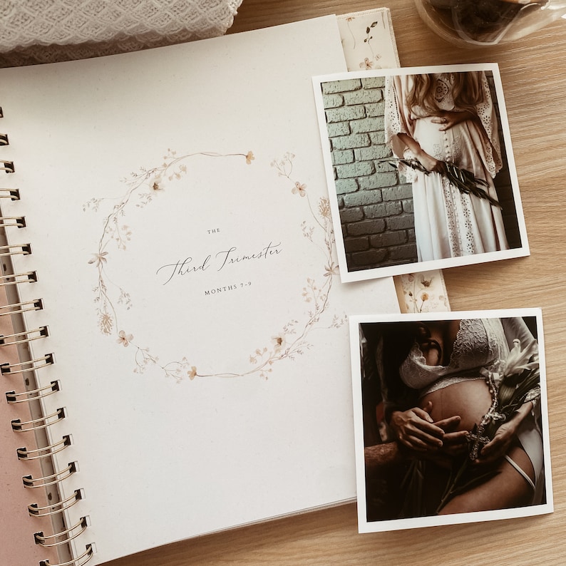 May include: A white journal with a gold spiral binding. The cover features a watercolor illustration of a floral wreath with the text "The Third Trimester Months 7-9". Two black and white photos of pregnant women are attached to the journal.