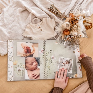 K&ouml;nnte beinhalten: Ein wei&szlig;es Babyalbum mit floralem Design und Fotos eines Babys. Das Album ist auf einer Seite ge&ouml;ffnet, die ein Foto von Babyf&uuml;&szlig;en, ein Foto eines schlafenden Babys und ein Foto eines Babys in einem wei&szlig;en Strampler zeigt.