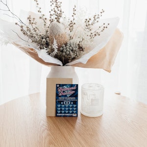 Pu&ograve; includere: Un mazzo di fiori secchi in un vaso bianco, una busta marrone e un bicchiere bianco con un disegno a linea di un viso. La busta ha un biglietto della lotteria gratta e vinci che dice "Diamond Dazzler Vinci fino a 100.000 &euro;".
