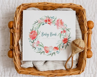 May include: A white baby book with a watercolor floral wreath design and the text "Baby Book Your First Years" in a wicker basket with a wooden rattle.