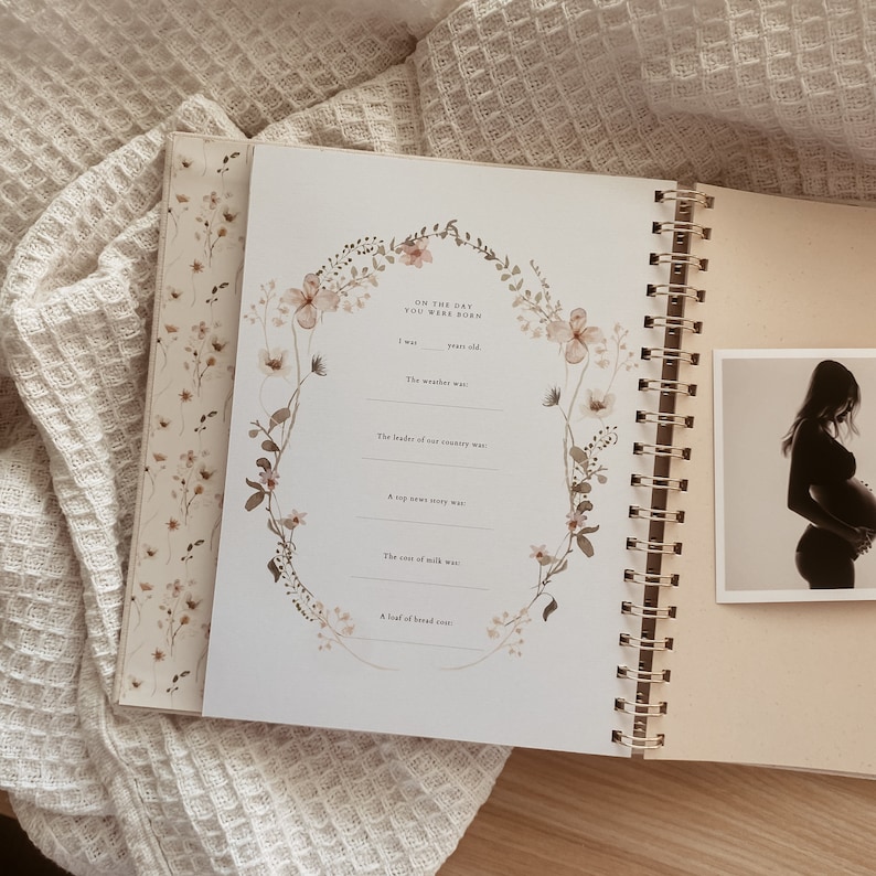 May include: A floral-themed journal with a gold spiral binding. The journal has a page with prompts for recording memories of the day a baby was born. The page includes prompts for the weather, the leader of the country, a top news story, the cost of milk, and a loaf of bread.