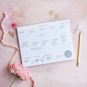May include: A white weekly planner with a pink background. The planner has sections for each day of the week, with handwritten notes. A pencil, paperclip, and decorative items surround the planner. The planner includes the text "Weekly Planner".