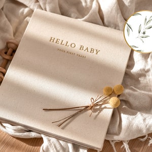 May include: A beige photo album with the text "HELLO BABY YOUR FIRST YEARS" in gold lettering. The album is lying on a beige fabric surface with a wooden toy and dried flowers.