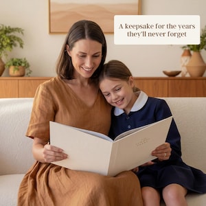 May include: A woman and a girl are seated on a white sofa, reading a book together. The woman wears a brown dress, and the girl wears a navy blue dress with a white collar. The book is open, with the text "A keepsake for the years they'll never forget".