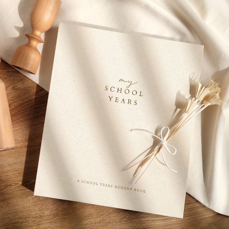 May include: A cream-colored memory book titled "my SCHOOL YEARS" with gold lettering. The book is adorned with a bundle of dried flowers tied with white string. The phrase "A SCHOOL YEARS MEMORY BOOK" is printed at the bottom.