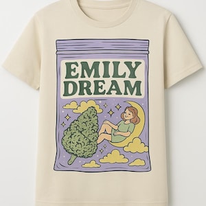 May include: A beige t-shirt featuring a graphic print of a purple bag with the text "EMILY DREAM" in green letters. The bag contains a green marijuana leaf and a cartoon woman sitting on a crescent moon.