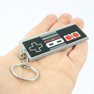May include: A gray and black Nintendo Entertainment System (NES) controller keychain with red buttons. The keychain is attached to a silver ring.