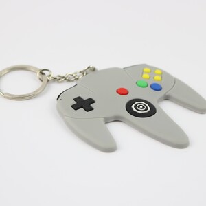 May include: A gray plastic keychain shaped like a Nintendo 64 controller. The controller has black buttons and a black and white control stick.
