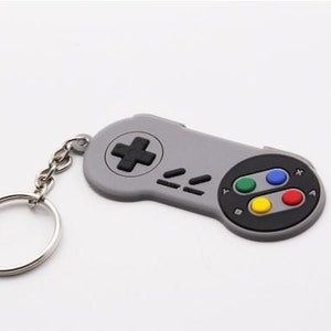 Può includere: Un portachiavi in plastica grigia a forma di controller Super Nintendo con pulsanti neri e pulsanti colorati. Il portachiavi ha un anello in metallo argentato.