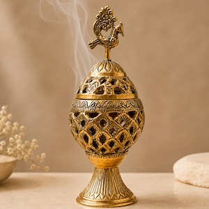May include: A gold-toned metal incense burner with an egg-shaped body featuring intricate cut-out patterns and a peacock-shaped finial on the lid. Wisps of white smoke rise from the top of the vessel, which stands on a flared pedestal base.