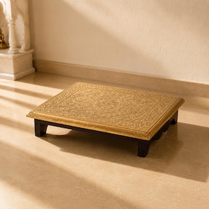 May include: A rectangular low platform with a metallic gold-coloured top surface featuring an intricate embossed floral and geometric pattern. The platform is supported by four short, dark-coloured wooden legs with a scalloped edge design.