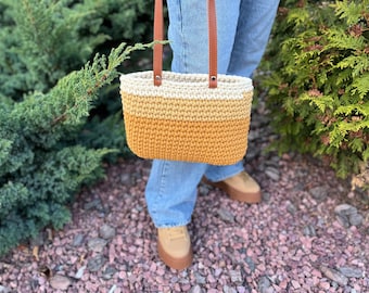 May include: A crocheted tote bag with a white and yellow ombre design. The bag has brown leather straps and is being held by a person wearing blue jeans.