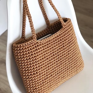 May include: A handcrafted, light brown macrame tote bag with braided handles. The bag has a rectangular shape and a textured, woven appearance. The bag is sitting on a white chair.