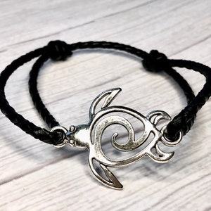May include: A black leather braided bracelet with a silver turtle charm. The turtle is a stylized design with a swirl in the center.