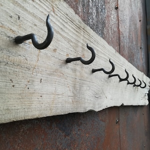 Puede incluir: Una fila de ganchos de metal rústico montados en una tabla de madera desgastada. Los ganchos son todos del mismo tamaño y forma, con un diseño curvo simple. La tabla está unida a una pared de metal oxidado.