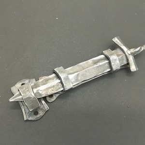 Puede incluir: Un pestillo de puerta de metal plateado con forma de espada con un mango decorativo. El pestillo está montado en una pared.