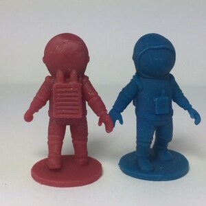 May include: Two astronaut figurines, one red and one blue, standing on circular bases. The astronauts are wearing spacesuits with backpacks and helmets.