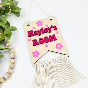 May include: A wooden sign with the text "Hayley's Room" in pink letters. The sign is decorated with pink flowers and has a white fringe hanging from the bottom.