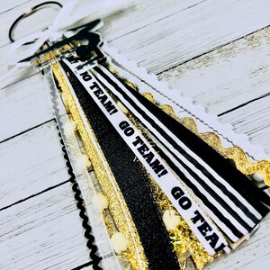 May include: Black and white striped ribbon with gold glitter ribbon and white pom poms. The ribbon has the text "Go Team! Go Team! Go Team!" printed on it.