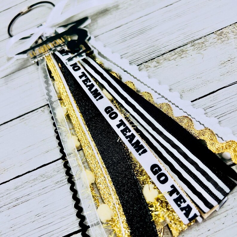 May include: Black and white striped ribbon with gold glitter ribbon and white pom poms. The ribbon has the text "Go Team! Go Team! Go Team!" printed on it.