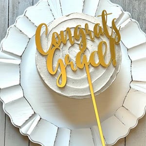 May include: A gold-colored acrylic cake topper featuring the script text "Congrats Grad!" with an exclamation point. The topper has a single vertical stake extending downward, designed to be inserted into the top of a round frosted cake.