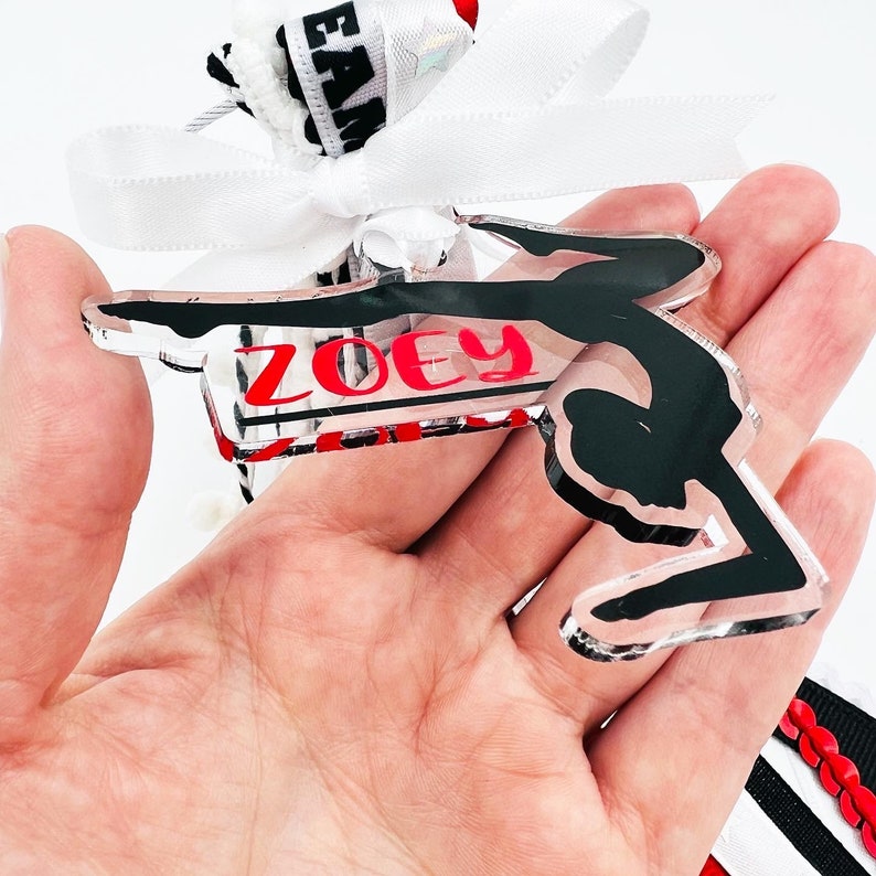 May include: A clear acrylic keychain with a black silhouette of a gymnast performing a handstand on the uneven bars. The keychain has the name "ZOEY" printed in red letters.