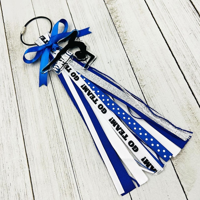May include: A blue and white cheer bow with a silver glitter ribbon and a black and white "Go Team" ribbon. The bow is attached to a black ring.