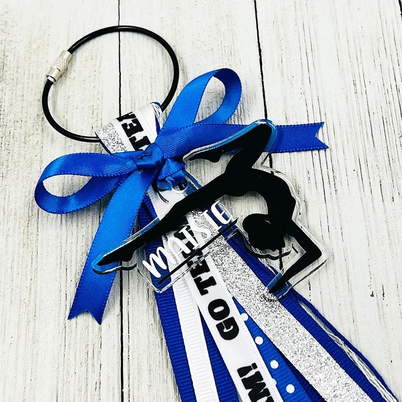 May include: A blue and silver cheer bow with a clear acrylic charm featuring a gymnast silhouette and the word "MARIS" on it. The charm is attached to a silver key ring.