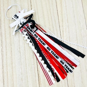 May include: A red, white, and black cheer bow with a black silhouette of a gymnast and the text "Go Team!" repeated on the ribbons. The bow is decorated with white pom-poms.