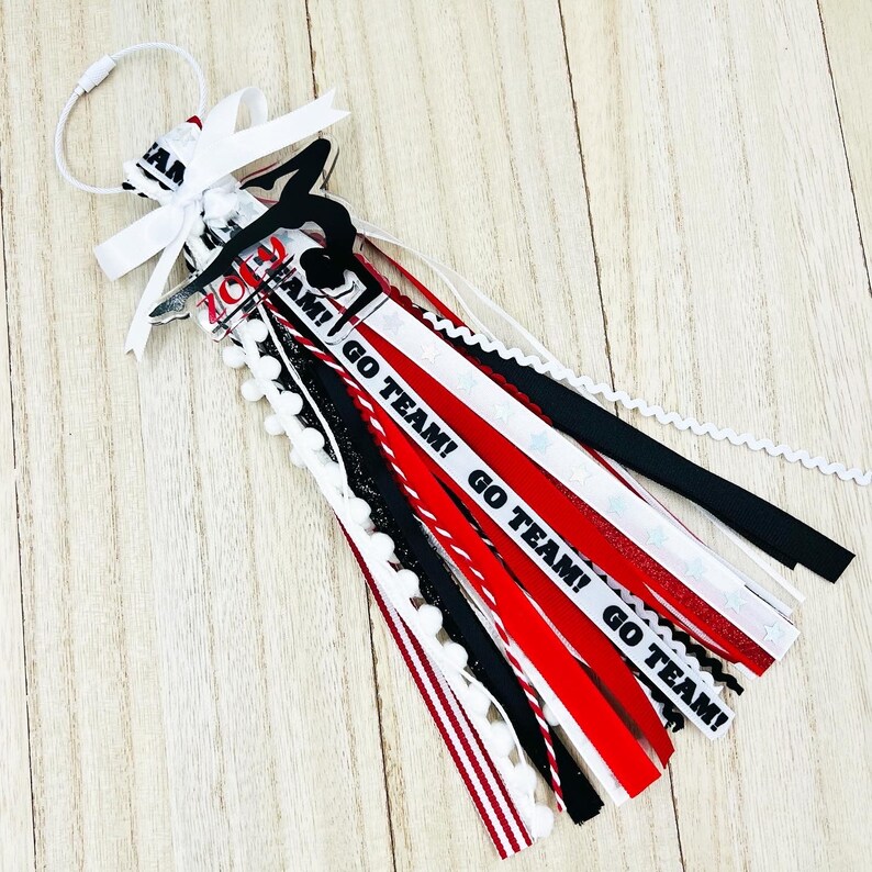 May include: A red, white, and black cheer bow with a black silhouette of a gymnast and the text "Go Team!" repeated on the ribbons. The bow is decorated with white pom-poms.