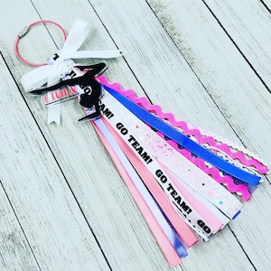 May include: A pink, white, and blue hair tie with a black silhouette of a gymnast on a bar. The hair tie has ribbons with the text "Go Team!" and "Go Team! Go" printed on them.
