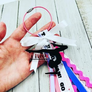 May include: A pink and white ribbon with the words "Go Team!" and "I'm with you" printed on it. A black silhouette of a gymnast on a bar is attached to the ribbon. The ribbon is attached to a silver keyring.