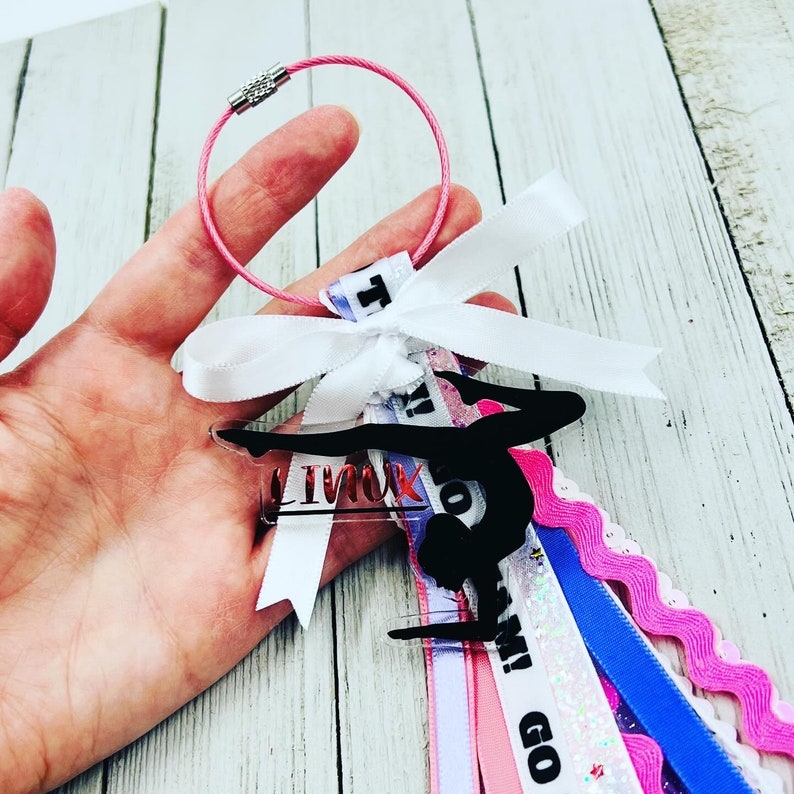 May include: A pink and white ribbon with the words "Go Team!" and "I'm with you" printed on it. A black silhouette of a gymnast on a bar is attached to the ribbon. The ribbon is attached to a silver keyring.