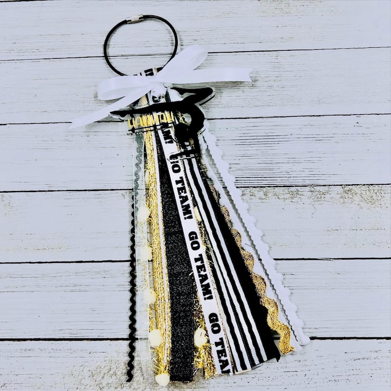 May include: Black and white cheer bow with gold glitter and white pom poms. The bow has a black and white striped ribbon with the text "GO TEAM! GO TEAM! GO TEAM!" printed on it.