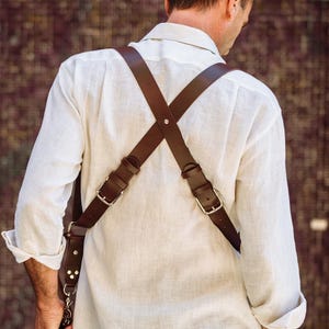 May include: A person wearing a white linen shirt with a brown leather camera strap. The strap has metal buckles and is worn across the chest and back.
