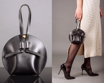 May include: A black leather handbag with a circular shape and a top handle. The bag has a gold-coloured button closure and decorative leather straps. The bag is held by a person wearing a cream dress and black high heels.