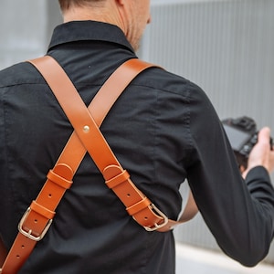 May include: A brown leather camera harness with gold-colored buckles. The harness crosses in an X-shape across the back and is worn over a black shirt. The image shows the back of a person wearing the harness.
