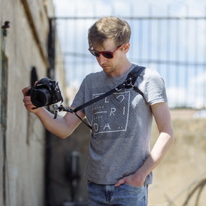 May include: A person wearing sunglasses and a grey t-shirt with the text "FLORIDA" printed on it, holding a black camera with a black strap.